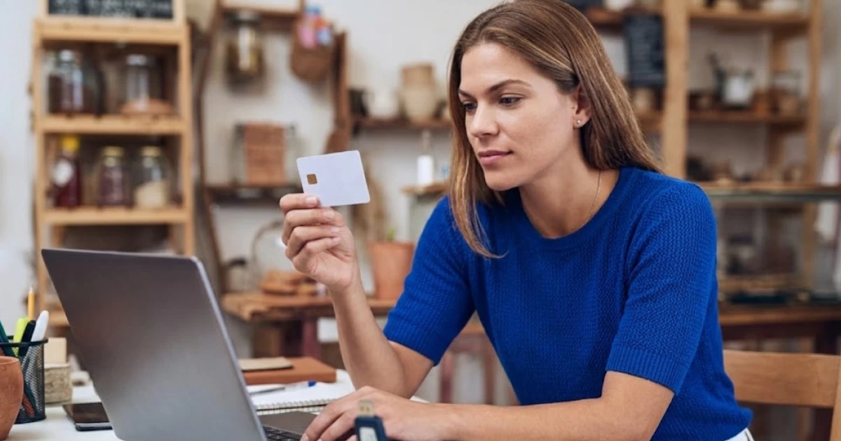 Empreendedora de pequeno negócio usa o notebook no balcão da loja segurando um cartão, com maquininha de cartão e calculadora sobre a mesa, cenário de emissão de nota fiscal com certificado digital.