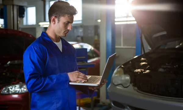 Como formar o preço de venda de serviços (mão de obra + custos) Profissional de mecânica de pé ao lado de um carro em manutenção na oficina usando um notebook para saber Como formar o preço de venda de serviços (mão de obra + custos).