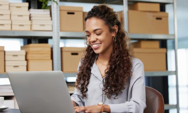 Como emitir nota fiscal para vendas em marketplace (Mercado Livre, Shopee, Magalu) Empreendedora trabalhando em seu notebook sobre uma mesa em ambiente de almoxarifado sobre Como emitir nota fiscal para vendas em marketplace (Mercado Livre, Shopee, Magalu).