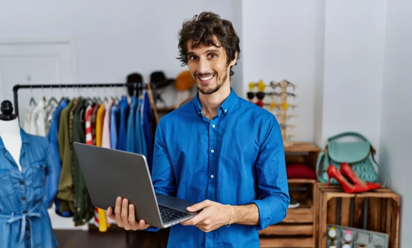 Planejamento fiscal de fim de ano para microempresa: tudo que o contador faz e você precisa saber Gerente de loja de roupas usando notebook, em pé no salão de vendas da loja representando: Planejamento fiscal de fim de ano para microempresa: tudo que o contador faz e você precisa saber.