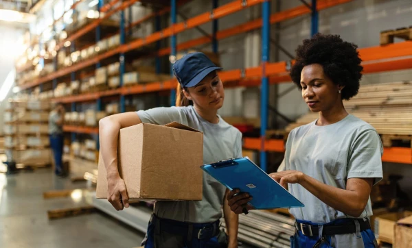 Gestão de fornecedores para distribuidora: como selecionar, negociar prazo e evitar ruptura de estoque Duas funcionárias femininas conversando sobre tarefas no estoque da empresa onde trabalham, uma com uma caixa na mão e outra com uma prancheta, representando: estão de fornecedores para distribuidora: como selecionar, negociar prazo e evitar ruptura de estoque