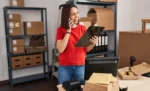 CNAE para distribuidora: como escolher o código correto e o impacto no regime tributário Funcionária de pé atendendo celular e com prancheta em mãos em ambiente de estoque representando: CNAE para distribuidora: como escolher o código correto e o impacto no regime tributário