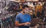 Sistema para bicicletaria: ordens de serviço, peças e controle financeiro Homem com notebook em mãos dentro de sua oficina de bicicletas usando o sistema para bicicletaria para gerenciar ordens de serviços, peças e controle financeiro