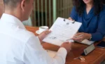 Alvará de funcionamento: o que é, como tirar e os riscos de operar sem ele homem com prancheta na mão sentado em uma mesa de frete a uma mulher representando: Alvará de funcionamento: o que é, como tirar e os riscos de operar sem ele