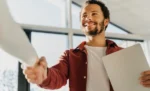 Como contratar o primeiro funcionário: passo a passo para MEI e ME Trabalhador sorridente com um documento em mãos dando um aperto de mão, representando: Como contratar o primeiro funcionário: passo a passo para MEI e ME