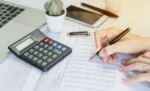 Folha de pagamento para pequenas empresas: o que compõe, como calcular e o papel do ERP Uma mão com uma caneta escrevendo em um documento ao lado de uma calculadora, representando: Folha de pagamento para pequenas empresas: o que compõe, como calcular e o papel do ERP