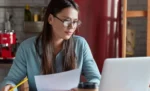 Como fazer análise de curva ABC de produtos no GestãoClick Mulher de óculos segurando um documento enquanto utiliza um notebook, representando a análise de curva ABC de produtos no Gestão Click 1x.
