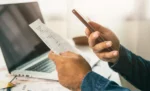 Como emitir boleto bancário pelo GestãoClick: passo a passo completo Mãos segurando um celular e um papel em frente a um notebook, representando: Como emitir boleto bancário pelo GestãoClick: passo a passo completo