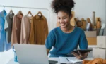 Integração ERP e marketplace: o que automatiza e como configurar no GestãoClick Proprietaria de loja de roupa sorrindo em frente ao seu notebook, representando: Integração ERP e marketplace: o que automatiza e como configurar no GestãoClick title: Integração ERP Marketplace: o que Automatiza?