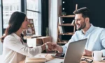 Como usar o upsell e cross-sell para aumentar a receita com a mesma base de clientes Em um escritório Mulher e homem apertam a mão como um gesto do que parece uma tratativa de um negócio, representando: Como usar o upsell e cross-sell para aumentar a receita com a mesma base de clientes